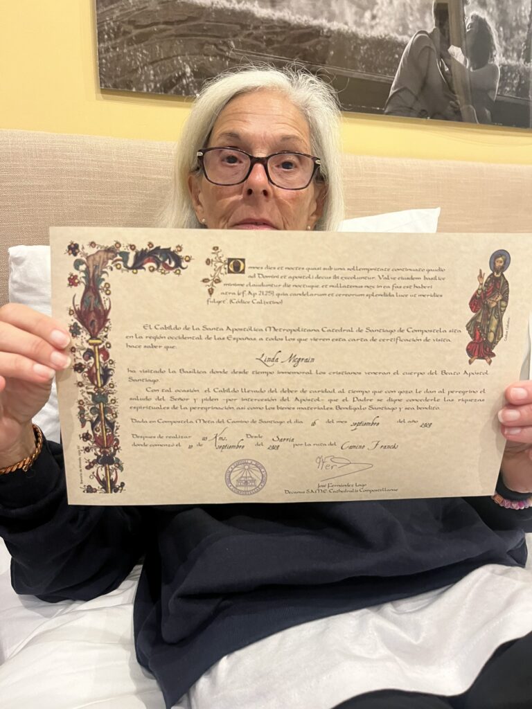 An older woman with silver hair and glasses proudly holding up a Compostela certificate, commemorating her completion of the Camino de Santiago pilgrimage. The certificate features ornate decorative borders and text in Spanish.