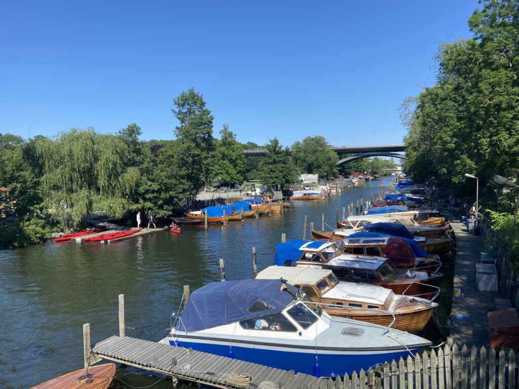 View of a canal lined with wooden boats covered with colorful tarps, bordered by lush green trees and a bridge in the background on a clear sunny day.