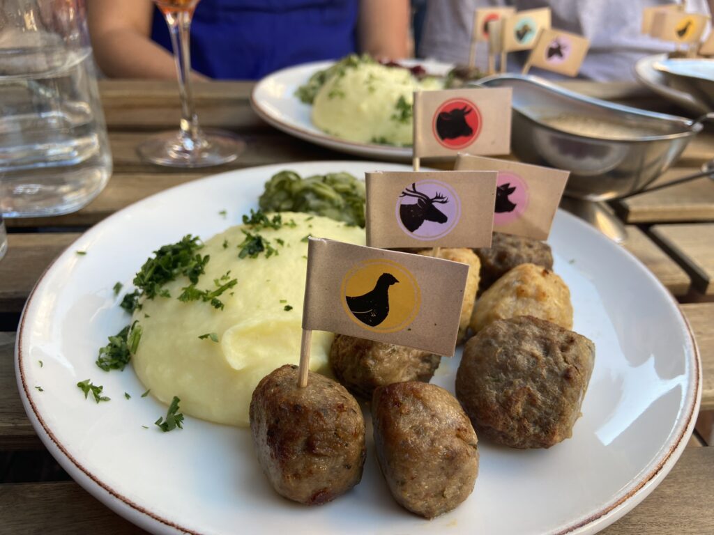 Plate of assorted meatballs served with mashed potatoes and greens, each meatball marked with a small flag indicating its meat type, such as reindeer, chicken, and beef, on an outdoor dining table.