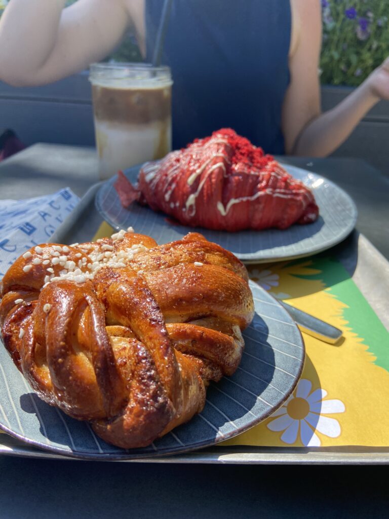 Close-up of a tray with a Swedish cinnamon bun topped with pearl sugar and a red velvet croissant drizzled with white icing, accompanied by an iced latte in the background.