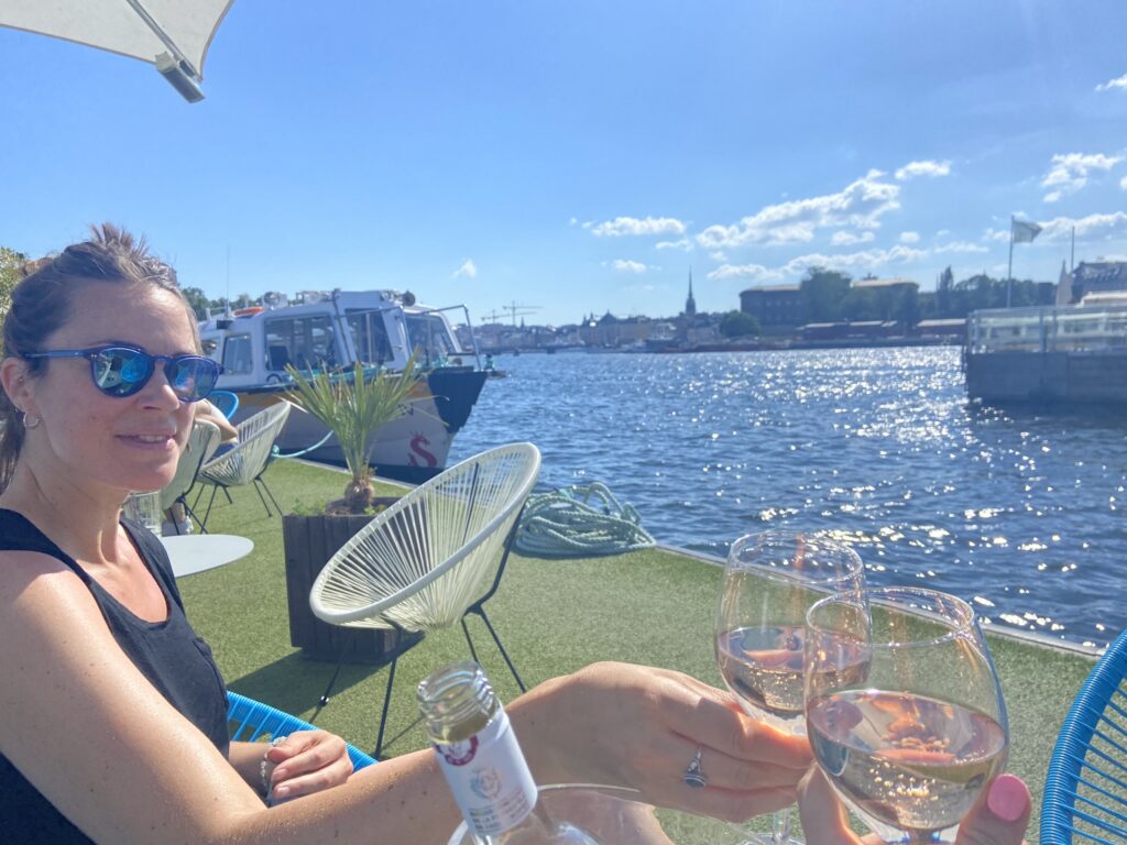 Person raising a glass of rosé while enjoying a sunny day by the water in Stockholm, with boats, blue sky, and city skyline in the background.