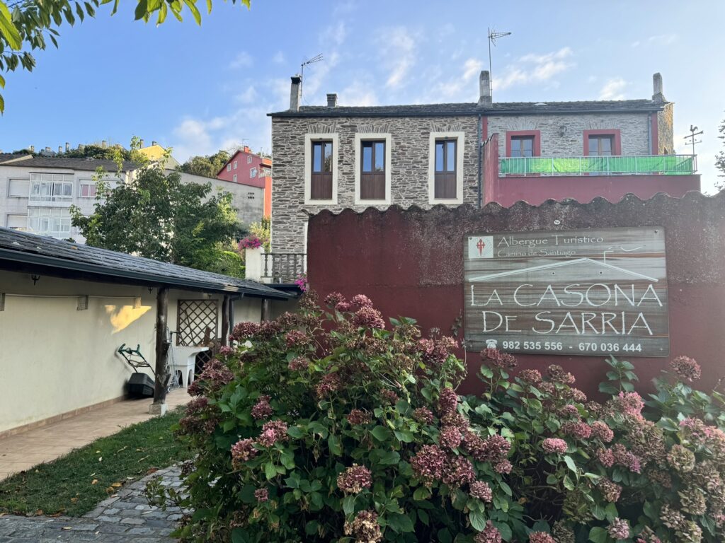 Exterior view of La Casona de Sarria, a tourist albergue on the Camino de Santiago. The rustic stone building is framed by blooming hydrangea bushes, with a wooden sign displaying the name and contact information for the accommodation.