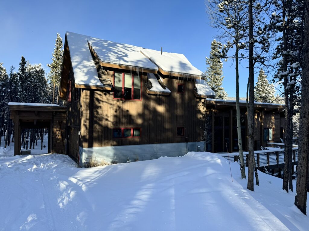 A rustic mountain cabin surrounded by snowy pine trees under a clear blue sky. The house features wooden siding, red-framed windows, and a snow-covered roof with icicles hanging from the eaves.