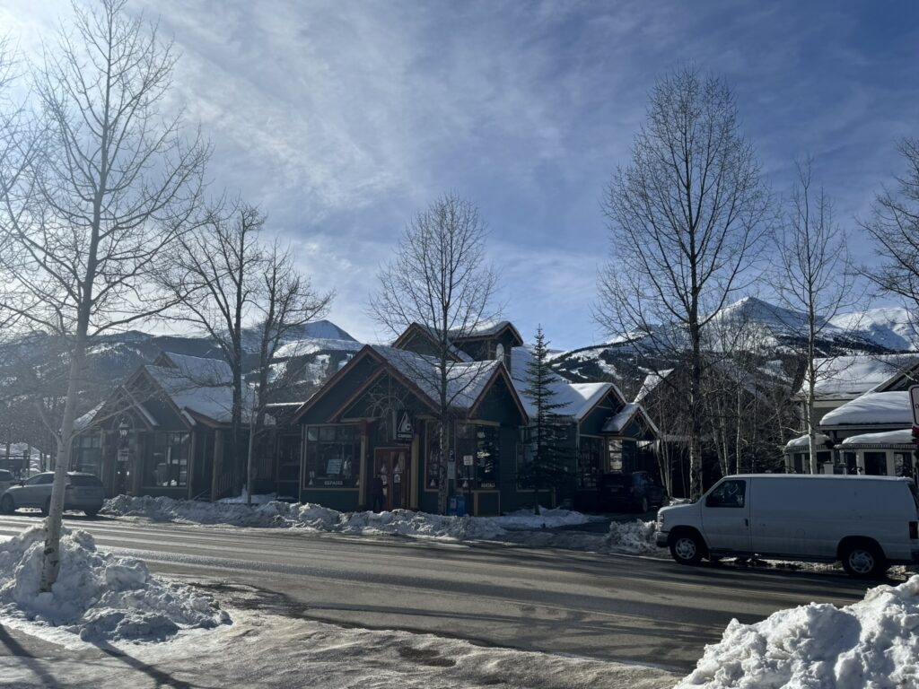 Snow-covered street in a mountain town with small shops, leafless trees, and snow-capped peaks in the background under a clear blue sky.
