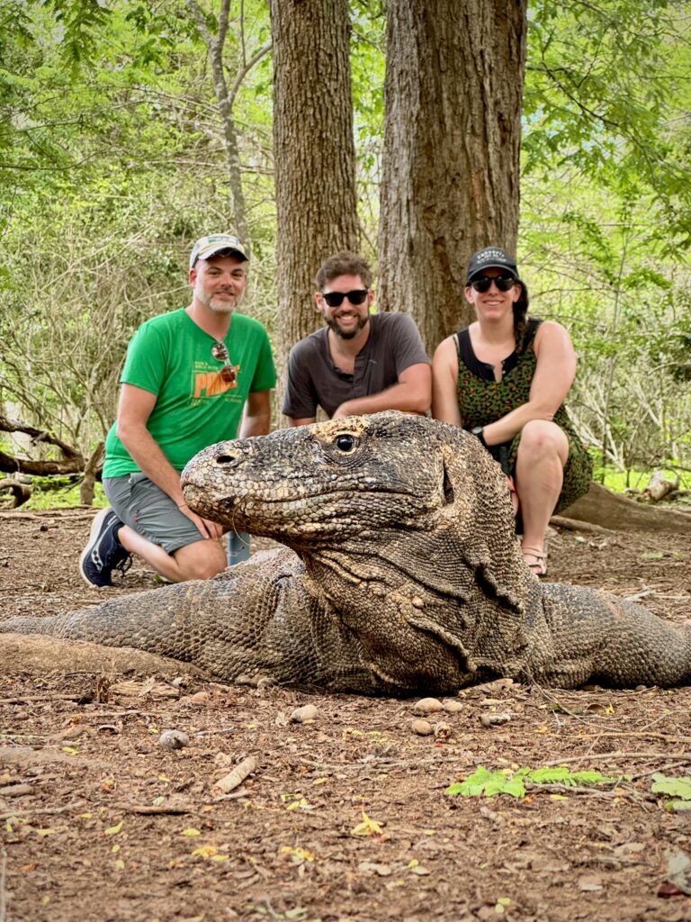 Three people pose behind a large Komodo dragon resting on the ground in a forested area, surrounded by tall trees and greenery.