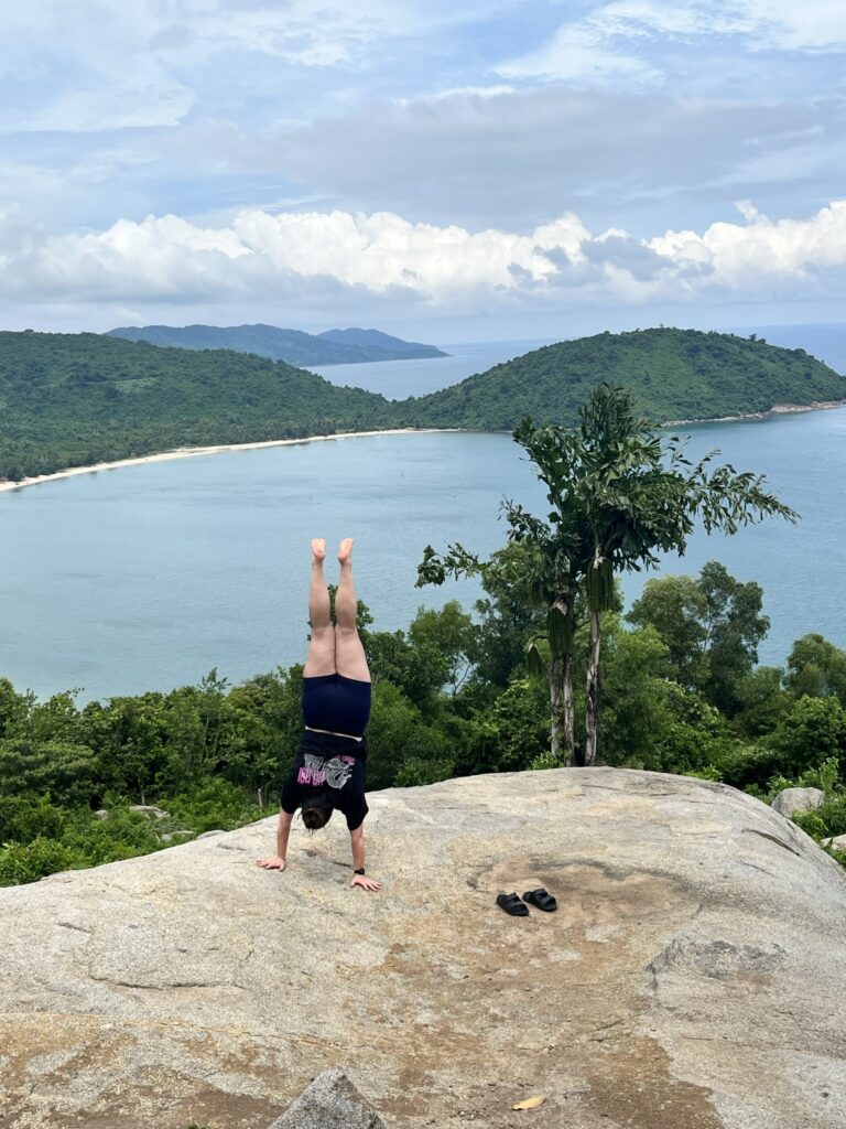 A person doing a handstand on a large rock overlooking a scenic bay surrounded by lush green hills and calm, blue water. The sky is partly cloudy, and a pair of sandals is placed on the rock nearby