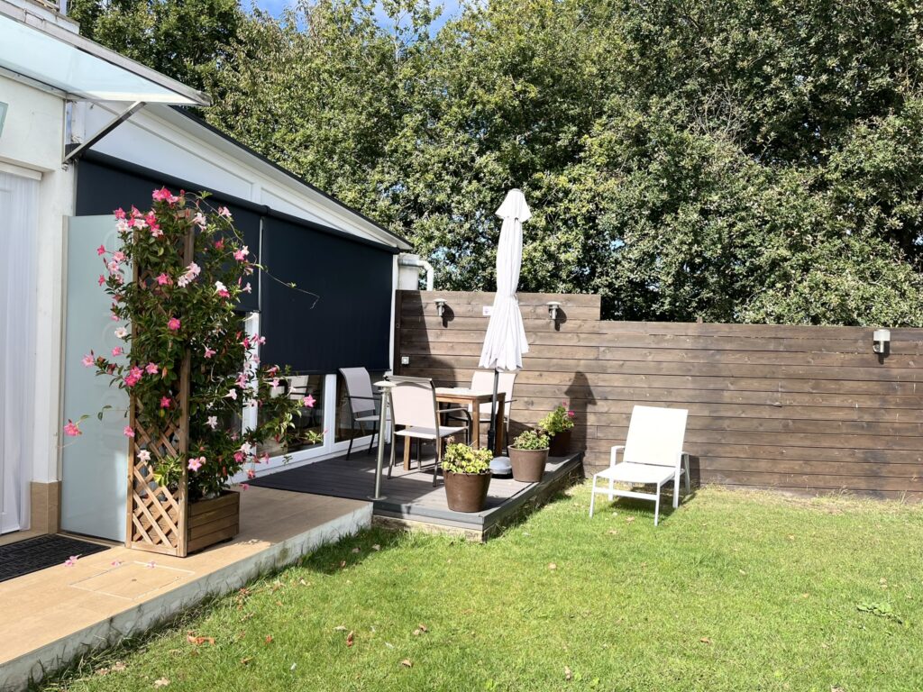 Cozy backyard patio with a wooden deck featuring an outdoor dining set and a white sun umbrella. Pink flowering vines climb a trellis near a shaded window, while a lounge chair sits on the grassy lawn next to a wooden privacy fence.