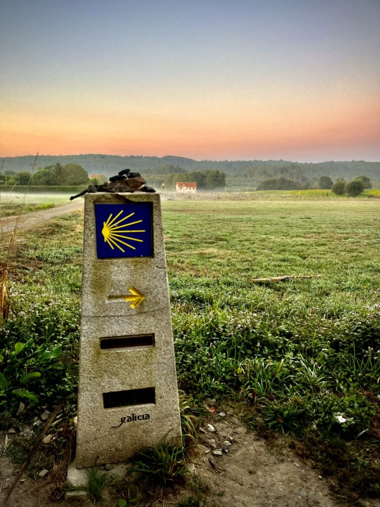 A stone Camino de Santiago marker with the blue and yellow scallop shell symbol and an arrow points the way along a grassy path at sunrise. The sky glows in soft pink and orange hues, with a distant house and tree-lined hills in the background.
